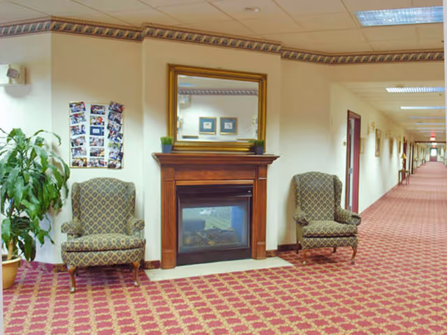 Carpeted senior living hallway seating area with two upholstered chairs flanking a fireplace topped by a mirror.