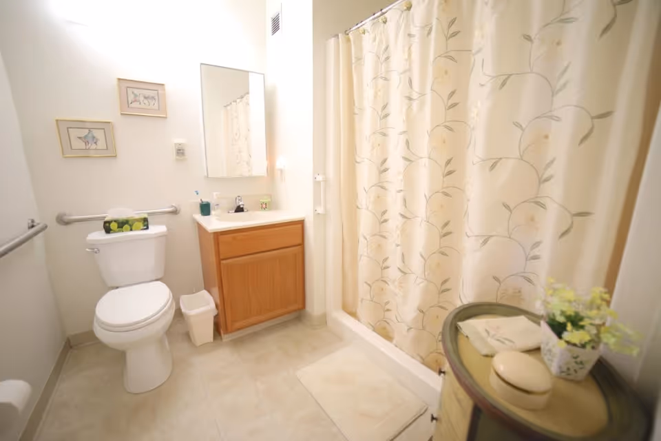 A clean and well-lit bathroom with a toilet, a wooden vanity with a sink and mirror above it, a shower with a floral-patterned curtain, and a small round table holding a potted plant and some toiletries.