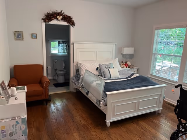 Bright bedroom with a white wooden bed, nightstand, orange armchair, window, and an open doorway revealing a bathroom.