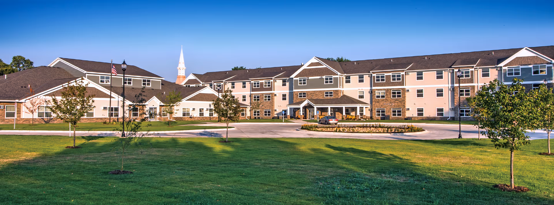 Front view of a large three-story senior living building with a circular driveway, landscaped lawn, and clear blue sky.