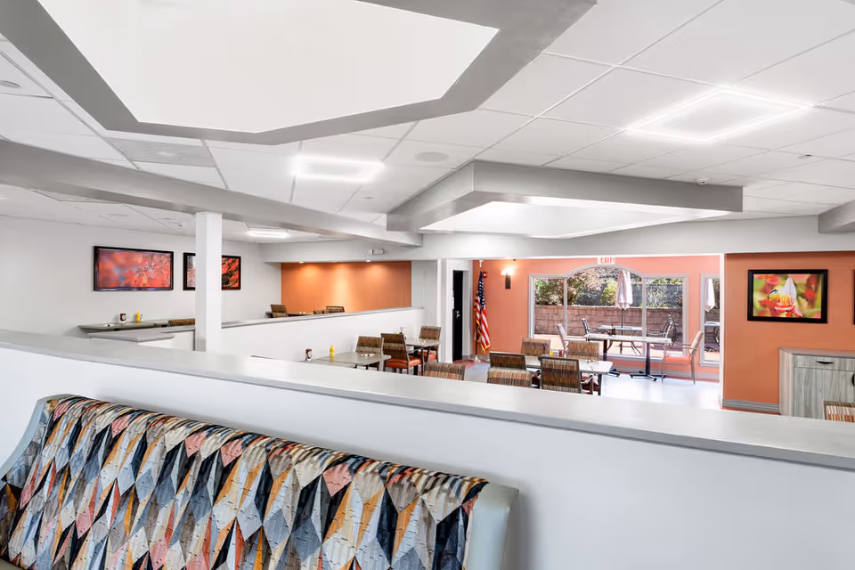 Interior view of a senior living facility dining area with multiple tables and chairs. The room has white walls with orange accent walls, framed floral artwork, and a large window showing an outdoor patio with tables and umbrellas. There is a patterned cushioned bench in the foreground and an American flag near the exit door.