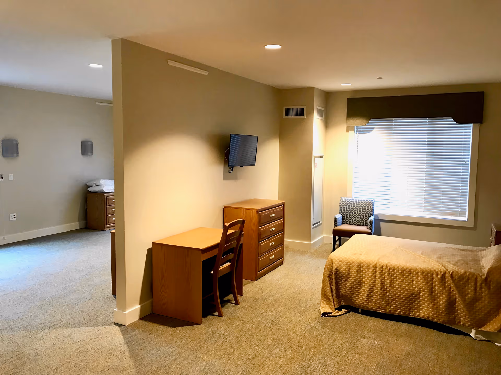 A senior living facility bedroom with a single bed covered in a beige quilt, a wooden desk and chair, a wooden dresser, a wall-mounted flat screen TV, a window with closed blinds and a valance, and a patterned armchair near the window. The room has beige walls and carpeted floor, with an adjacent open area visible to the left.