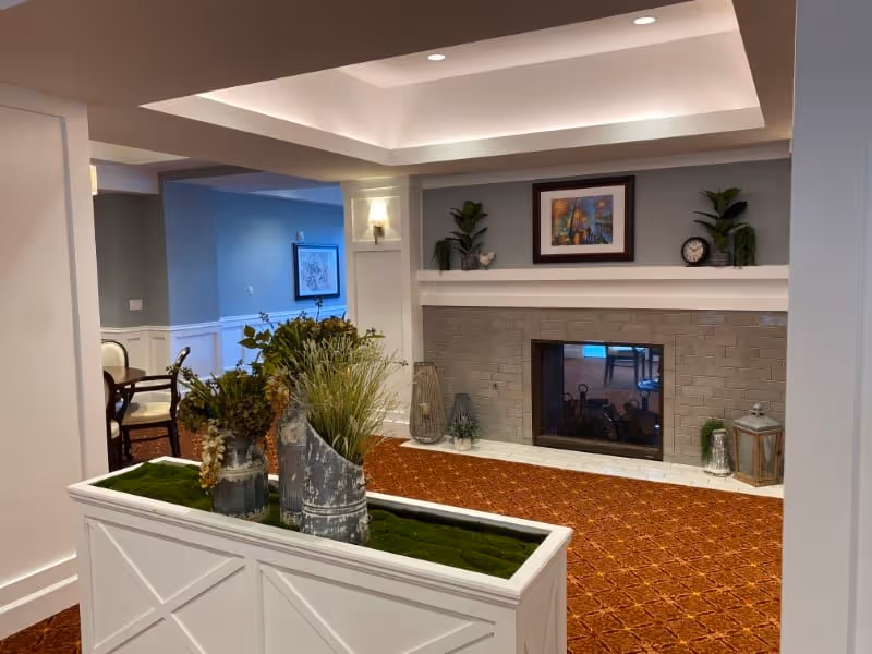 Interior view of a senior living facility lounge area with a decorative fireplace, plants, and framed artwork above the mantel. The room has a patterned carpet, recessed ceiling lighting, and a white planter with greenery in the foreground. A dining area with chairs and a framed picture is visible in the background.