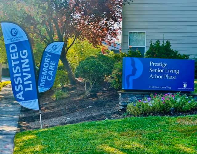 Outdoor landscaped entrance with a blue sign reading "Prestige Senior Living Arbor Place" and two vertical flags for Assisted Living and Memory Care.