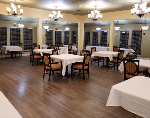 Spacious dining room with multiple tables covered in white tablecloths, upholstered chairs, chandeliers, and mirrored walls.