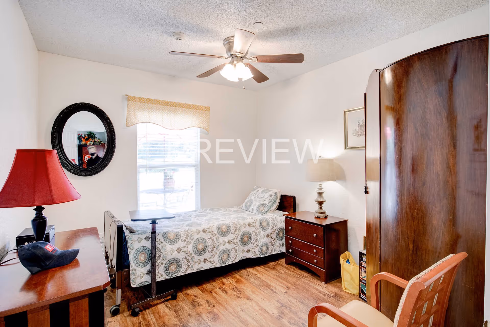A small bedroom with a single bed covered in a patterned bedspread, a wooden nightstand with a lamp, a wooden wardrobe, a wooden chair, a table with a red lamp and a black cap, a round mirror on the wall, and a window with blinds and a valance. The ceiling has a fan with lights, and the floor is wooden.