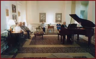 A group of people sitting in a communal lounge with sofas, a coffee table, and a grand piano.