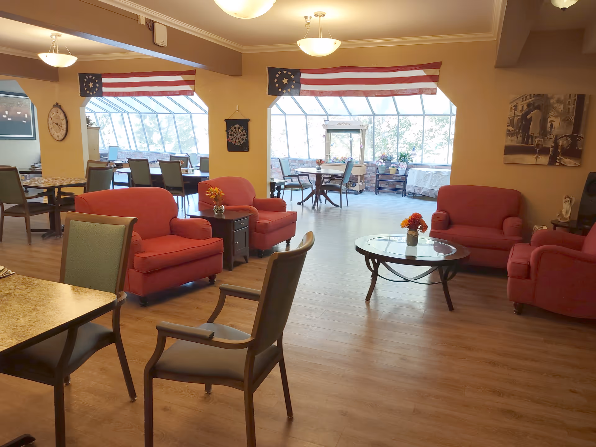 A spacious senior living common area with red upholstered armchairs and sofas arranged around a glass coffee table with a small flower vase. There are additional dining tables and chairs in the background near large windows that let in natural light. Two American flags hang above the windows, and a dartboard is mounted on the wall. The room has warm lighting and wooden flooring.