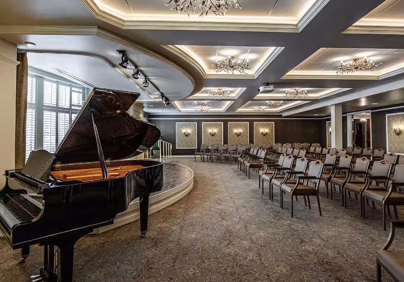 A spacious event room with a grand piano on a small stage to the left and rows of chairs arranged facing the stage. The room features elegant ceiling lighting fixtures, wall sconces, and large windows with white shutters allowing natural light to enter.