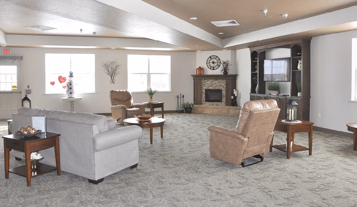A spacious living room area in an assisted living facility with a gray sofa, two brown armchairs, wooden side tables, a round coffee table, a fireplace with a clock above it, a large dark wood entertainment center with a TV, and large windows letting in natural light.