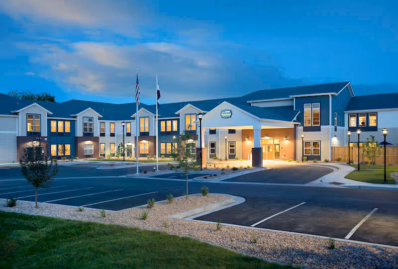 Front exterior of a two-story senior living facility at dusk with an illuminated entrance, flagpoles, and an empty parking area.