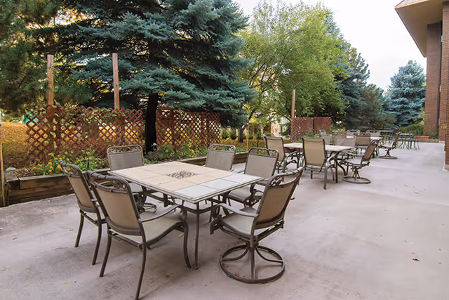 Outdoor patio area with multiple metal tables and chairs arranged on a concrete surface, surrounded by trees and wooden lattice fencing.