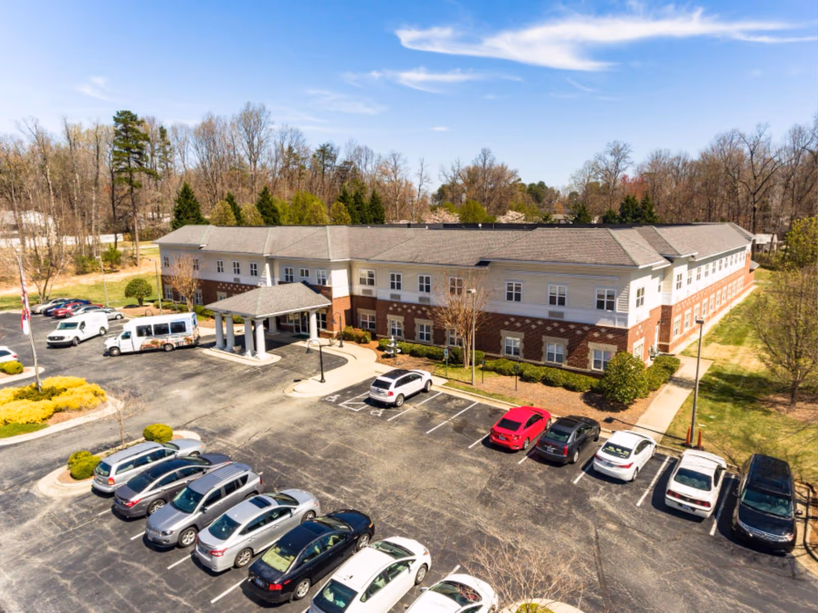 Aerial view of a two-story senior living facility building with a covered entrance and multiple parked cars in the parking lot. The building is surrounded by trees and greenery under a clear blue sky.