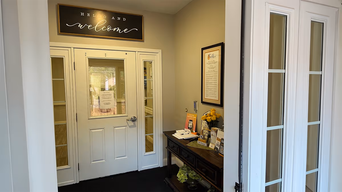 Interior entryway showing a white door with a 'welcome' sign above and a console table holding brochures and flowers.
