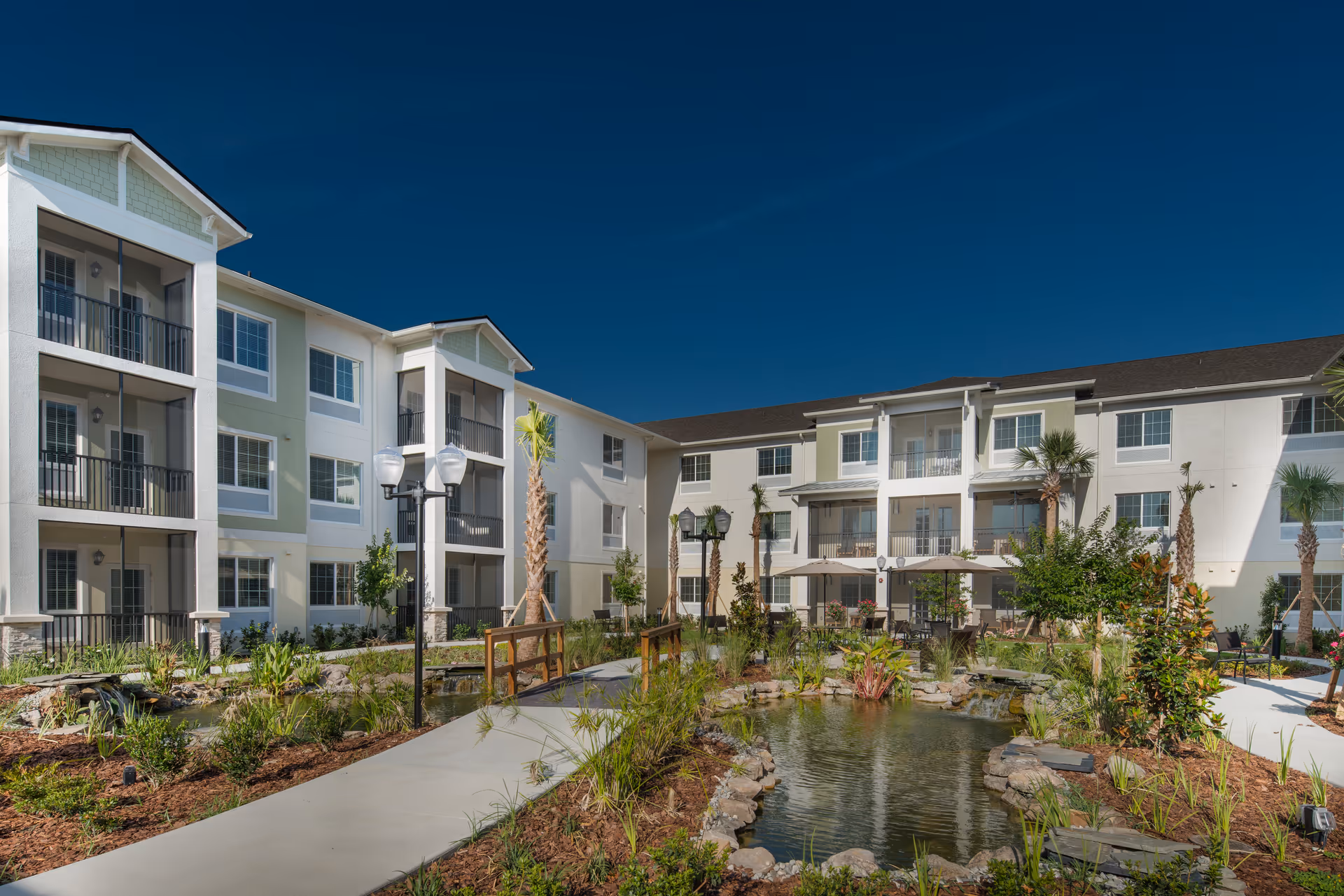 Outdoor courtyard area of a senior living facility with a pond surrounded by rocks and plants, paved walkways, palm trees, and a three-story building with balconies in the background under a clear blue sky.