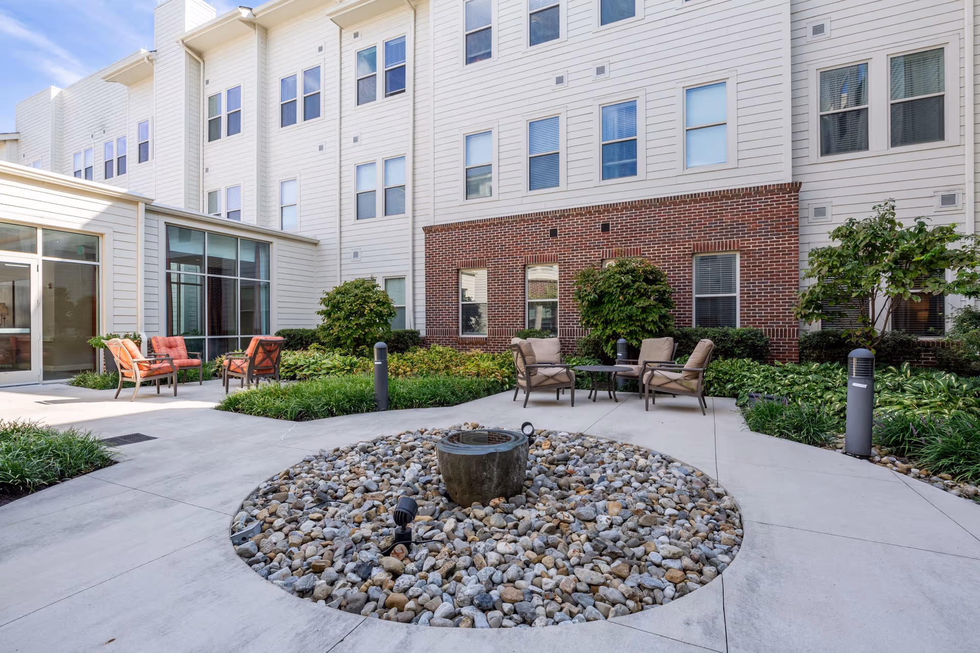 Outdoor courtyard area of a senior living facility with a circular rock garden featuring a small water fountain in the center. Surrounding the garden are concrete walkways and seating areas with cushioned chairs. The building exterior has white siding and red brick accents with multiple windows.