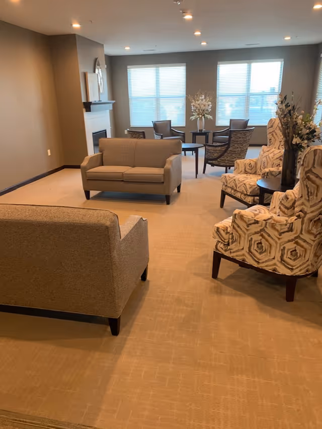 Spacious communal living room with sofas, patterned armchairs, small tables, and large windows.