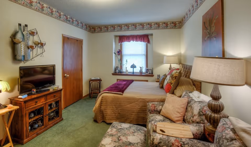A cozy bedroom in a senior living facility featuring a bed with a brown quilt and multiple pillows, a floral-patterned armchair with a wooden tray, a wooden TV stand with a flat-screen television, a window with a red valance, and decorative wall art including a metal wine bottle and glass sculpture and a floral painting. The room has green carpet and floral wallpaper border near the ceiling.