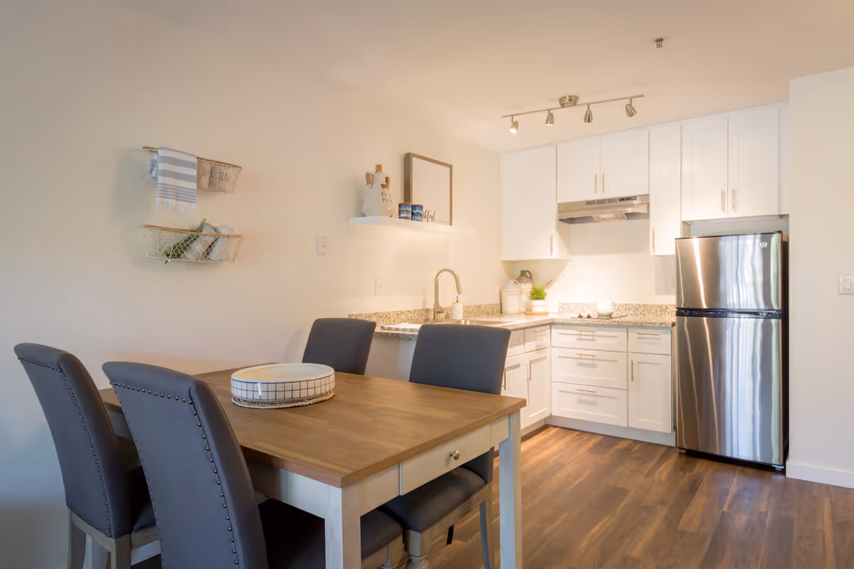 A modern kitchen and dining area with white cabinets, granite countertops, a stainless steel refrigerator, and a wooden dining table with four gray upholstered chairs. The kitchen has a track light fixture on the ceiling and decorative items on a floating shelf and countertop. The floor is wood with a warm tone.