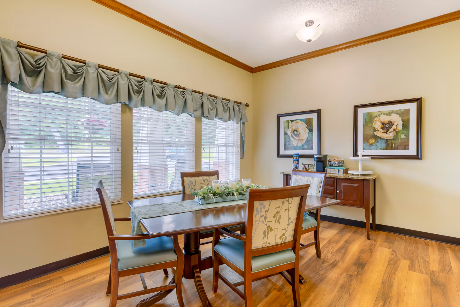 A dining room with a wooden table and four chairs with floral upholstery. The table has a green table runner and a centerpiece with candles and greenery. There are three large windows with blinds and green valances. On the wall, there are two framed floral paintings above a small wooden sideboard with a coffee maker and some decorative items. The floor is wooden, and the walls are painted light yellow with a wooden crown molding.