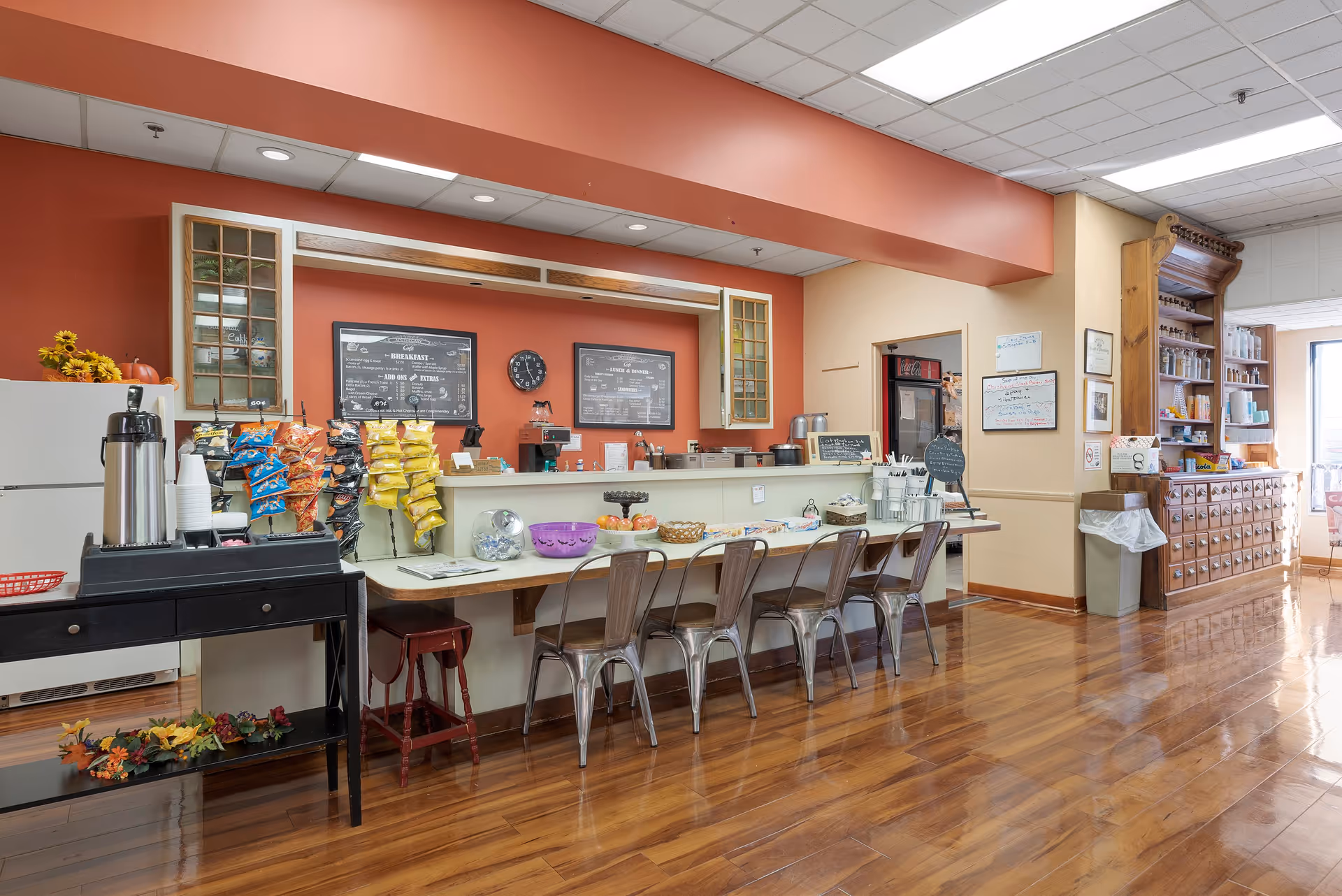 Interior view of a snack and beverage counter area in a retirement community with a row of metal chairs along a counter, a variety of chips displayed on racks, coffee dispensers, and a refrigerator. The room has wooden floors, warm-colored walls, and shelves stocked with various items.