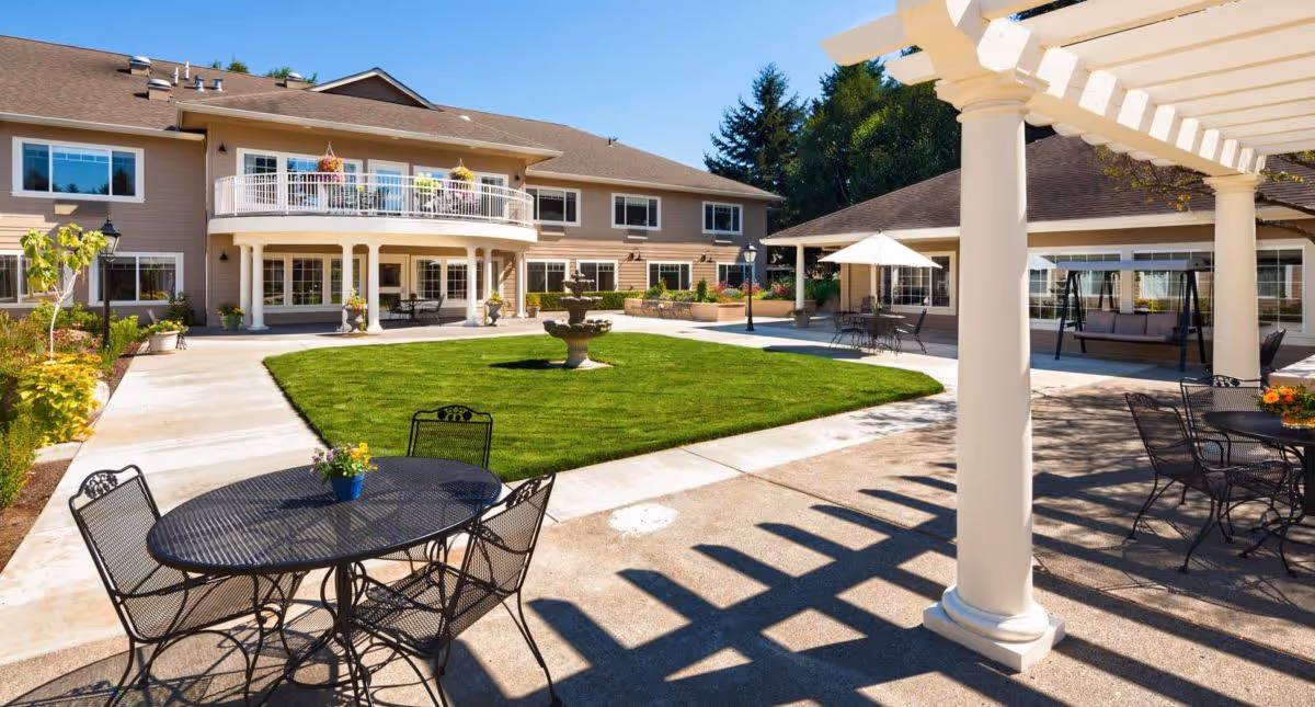 Outdoor courtyard area of The Hampton and Ashley Inn Memory Care Community featuring a green lawn with a central fountain, surrounded by paved walkways, black metal tables and chairs with flower pots, a white pergola casting shadows, and a two-story beige building with balconies and large windows under a clear blue sky.
