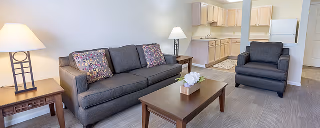 A bright living room area featuring a dark gray sofa with two patterned throw pillows, a matching armchair, two wooden side tables each with a lamp, and a wooden coffee table with a small flower arrangement. In the background, there is a kitchen area with light wood cabinets, a white refrigerator, and a sink.