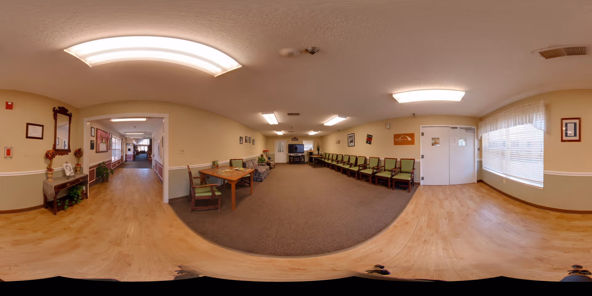 A spacious senior living facility common area with a row of green cushioned chairs along the right wall, a wooden table with chairs, a patterned sofa, and a TV on a stand at the far end. The room has beige walls with white trim, wood flooring near the windows, and carpet in the seating area. There are large windows with white blinds letting in natural light, framed pictures on the walls, and ceiling lights providing illumination. A hallway extends to the left side of the image with additional seating and decor.