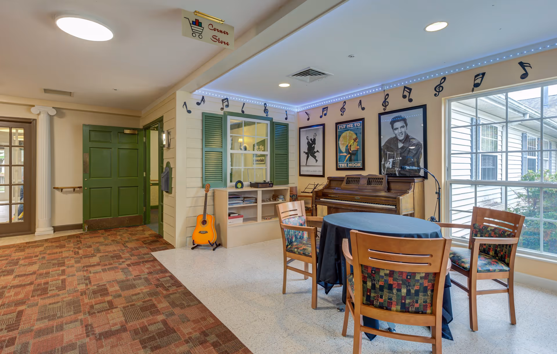 A cozy common area in a memory care community featuring a piano with a microphone, a guitar leaning against a small bookshelf, and a round table with four chairs. The walls are decorated with musical notes and framed posters, including one of Elvis Presley. There is a large window letting in natural light and a green door leading to another room. A sign above reads 'Corner Store.'