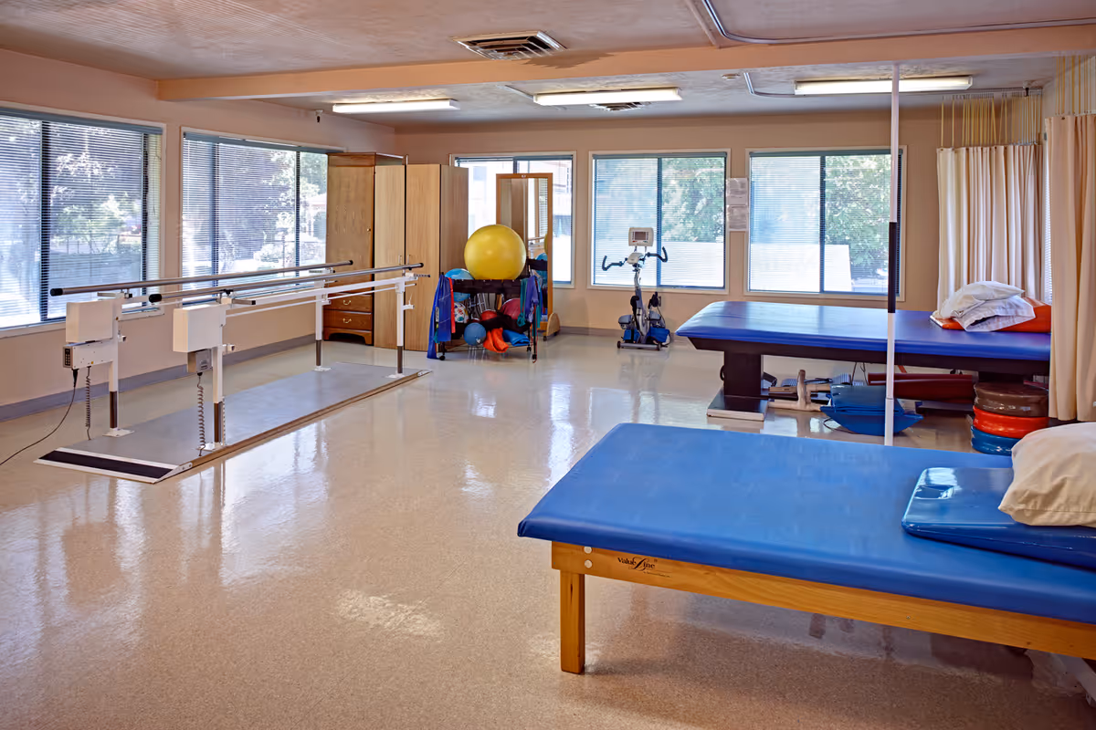 A spacious physical therapy room with large windows letting in natural light. The room contains parallel bars for walking exercises, two padded therapy tables with pillows, an exercise bike, a large yellow exercise ball, and various smaller exercise equipment. The floor is shiny and clean, and there are curtains for privacy around one therapy table.