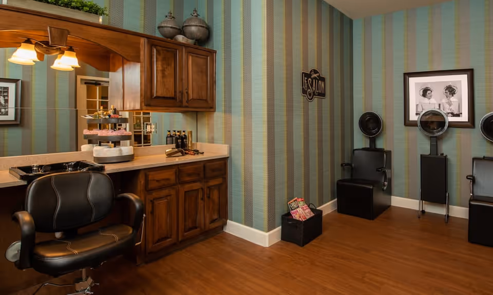 Interior of a salon area with striped wallpaper, wooden cabinets, a black salon chair in front of a counter with a mirror, and two black hair drying chairs against the wall. A framed black and white photo and a sign that reads 'Le Salon' are also visible.