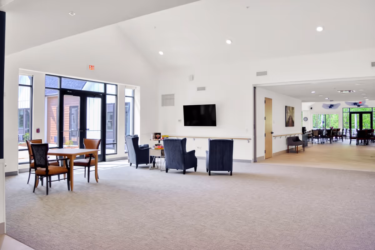 A bright and spacious common area in a senior living facility featuring a seating arrangement with four dark armchairs around a small table, a wall-mounted flat-screen TV, and a wooden table with two chairs near large glass doors leading outside. In the background, there is a dining area with multiple tables and chairs, large windows, and a door.