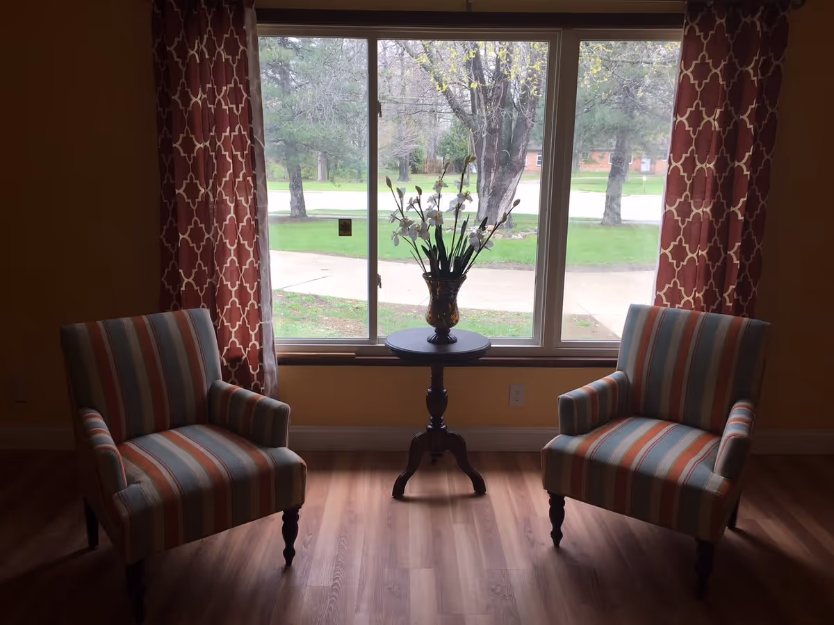 Two striped armchairs facing each other with a small round table in between, holding a vase with flowers, positioned in front of a large window with red patterned curtains. Outside the window, trees and a sidewalk are visible.