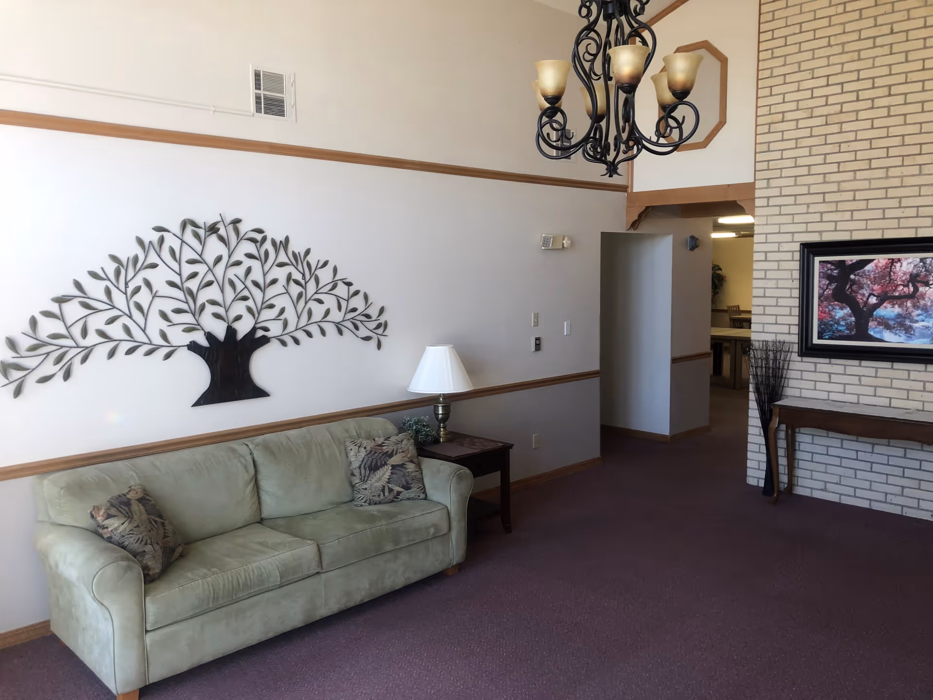 A living room area with a light green couch adorned with two patterned pillows, a wooden side table with a lamp, and a decorative metal tree wall art above the couch. There is a chandelier hanging from the ceiling, a framed picture of a tree on a brick wall, and a wooden console table beneath it. The room has beige walls with wooden trim and purple carpet flooring.