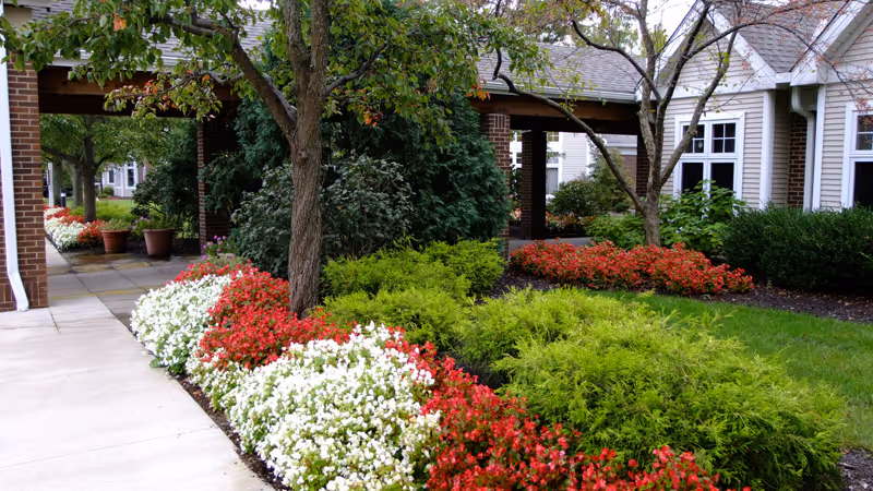 A landscaped outdoor area at Kendal at Oberlin featuring a walkway bordered by vibrant red and white flowers, green shrubs, and trees. The scene includes parts of a building with brick columns and white siding, along with large windows and a covered walkway.