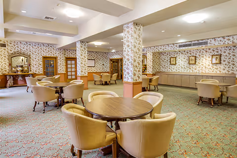 A spacious common area with round wooden tables surrounded by beige upholstered chairs. The room features floral wallpaper, carpeted flooring with a patterned design, and several framed pictures on the walls. There are also wooden doors and a sideboard with decorative mirrors and lamps.