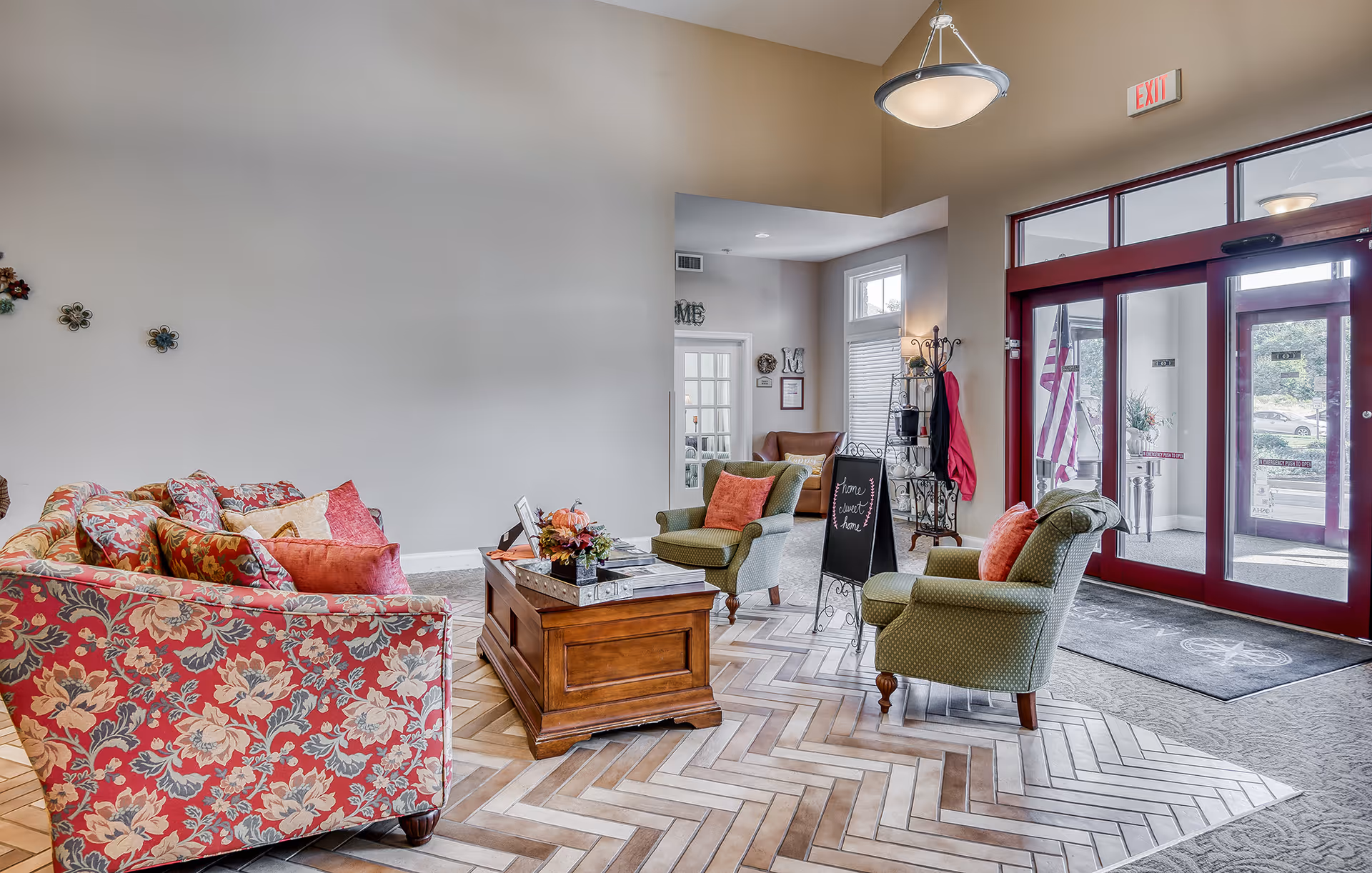 A bright and welcoming senior living facility lobby with floral patterned sofas, green armchairs with orange cushions, a wooden coffee table with decorative items, a chalkboard sign, and large glass doors with an exit sign above. The floor features a mix of carpet and herringbone patterned tiles, and there is an American flag visible outside the glass doors.