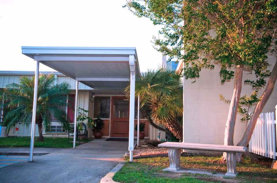 Entrance to a building with a covered walkway supported by metal poles, surrounded by palm trees and other greenery. A stone bench is positioned on the grass near the walkway, and the building exterior is light-colored with windows and a door visible.