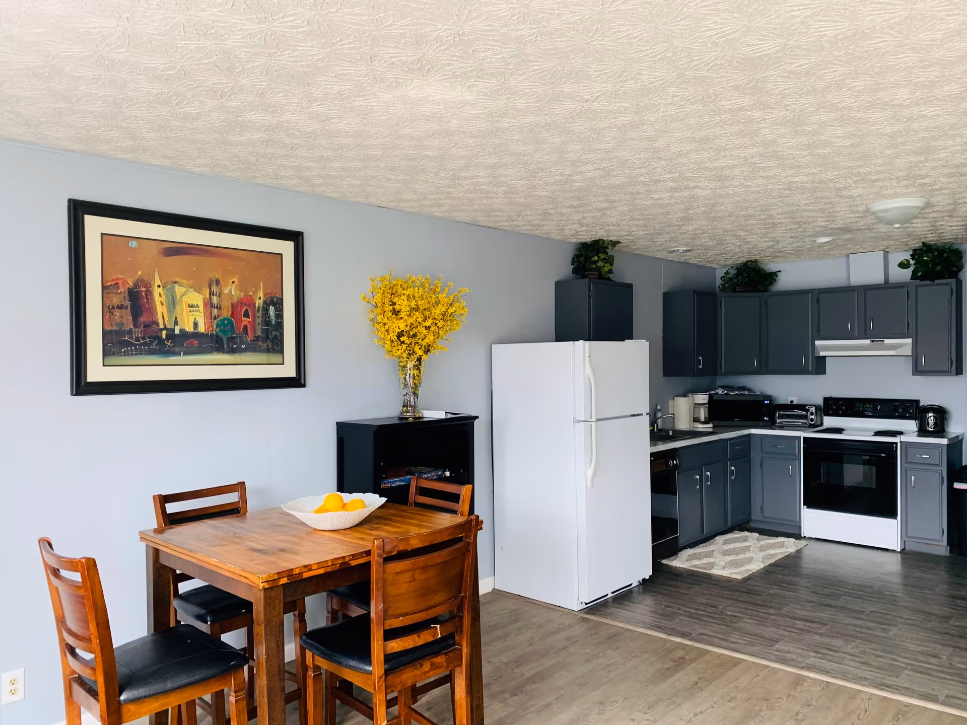 Open-plan kitchen and dining area with a wooden table and chairs, white refrigerator, gray cabinets, and framed wall art.