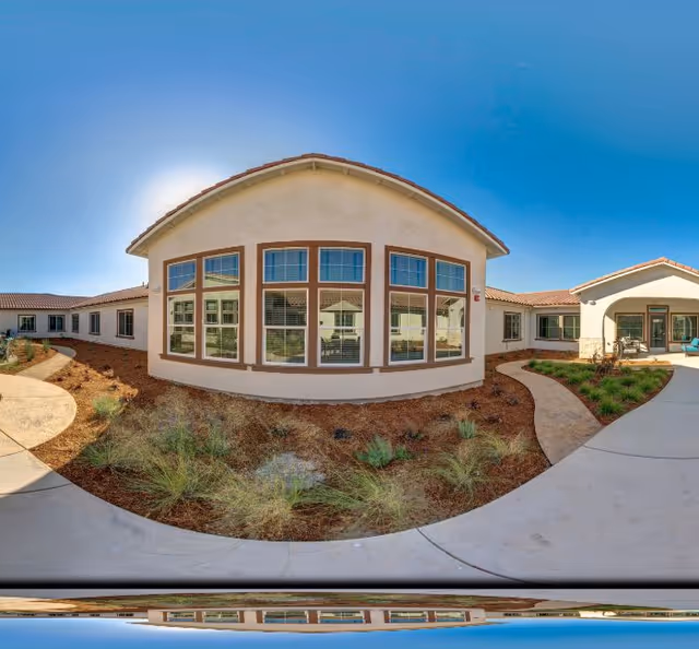 Exterior view of a single-story senior living building with large windows overlooking a landscaped courtyard and curved walkways under a clear blue sky.