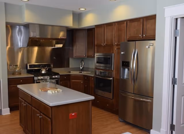 Modern kitchen with wooden cabinets, stainless steel refrigerator, oven, microwave, and stove. There is a kitchen island in the center with a glass cake dome and some bread on it. The floor is wooden and the walls are painted light gray.