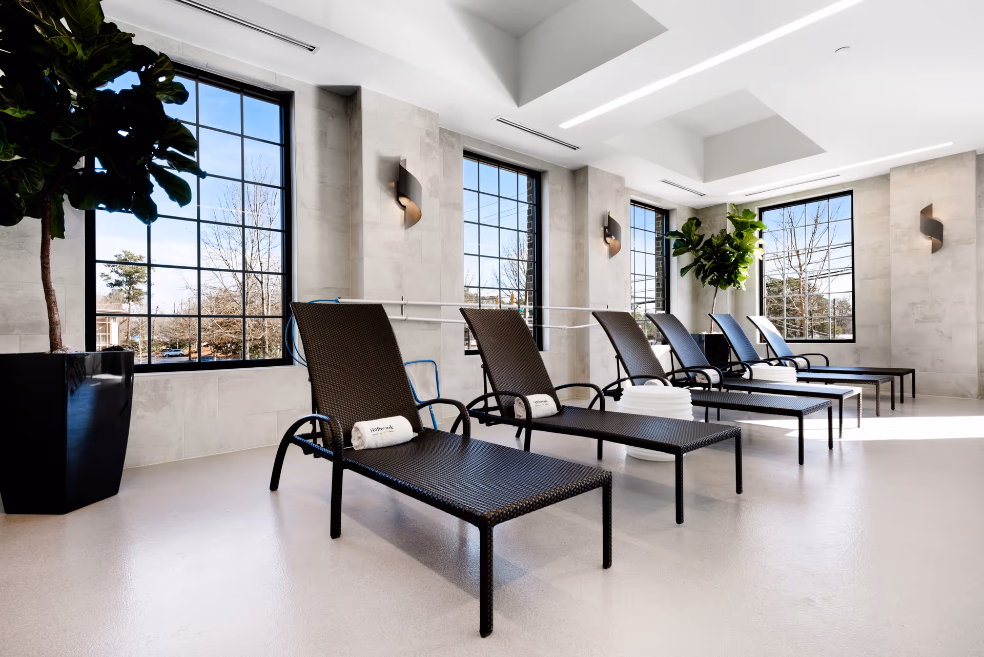 A bright indoor relaxation area with six black woven lounge chairs arranged in a row, each with a rolled white towel placed on it. Large windows with black frames let in natural light, and there are two large potted plants in the corners. The walls are light-colored with modern wall sconces, and the floor is a smooth, light surface.