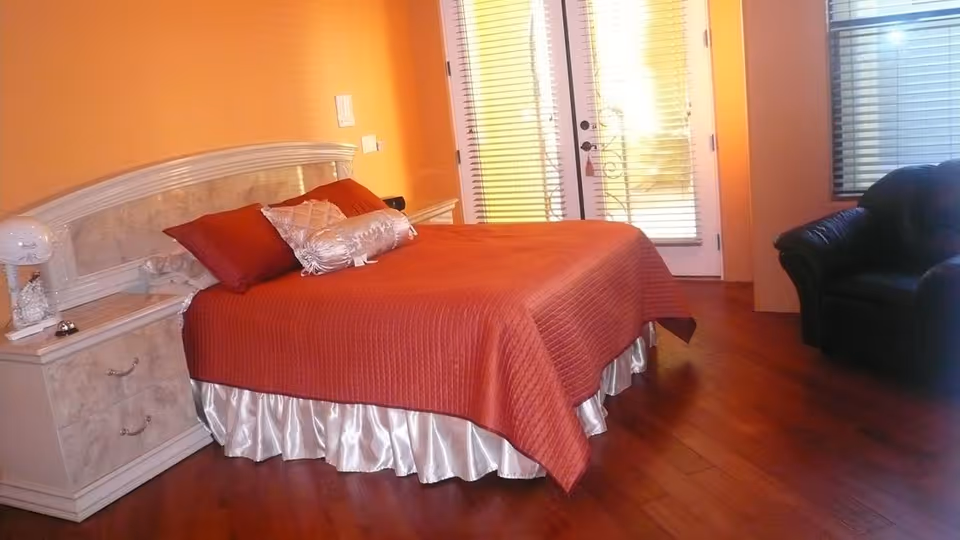 A bedroom with an orange bedspread on a bed, a white headboard, a nightstand with a lamp and a small bell, wooden flooring, a black armchair, and a door with window blinds leading outside.