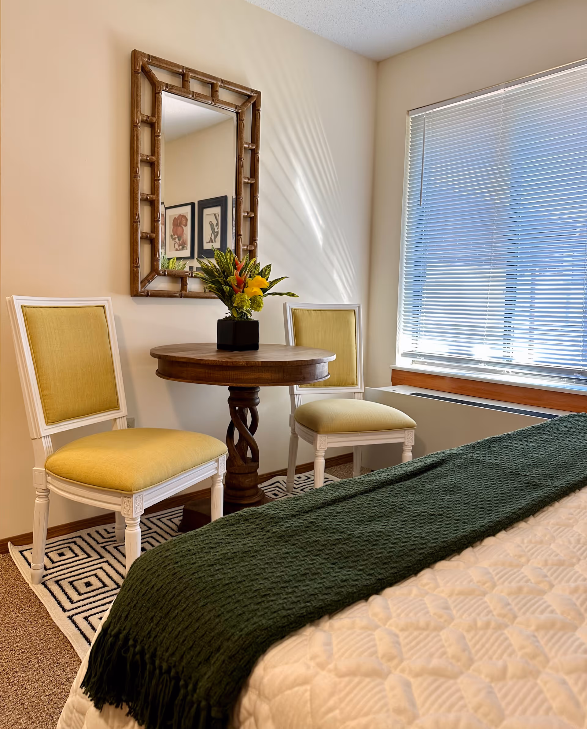 A cozy bedroom corner with a small round wooden table, two yellow-upholstered chairs, a mirror on the wall, a window with blinds, and a bed with a green throw.