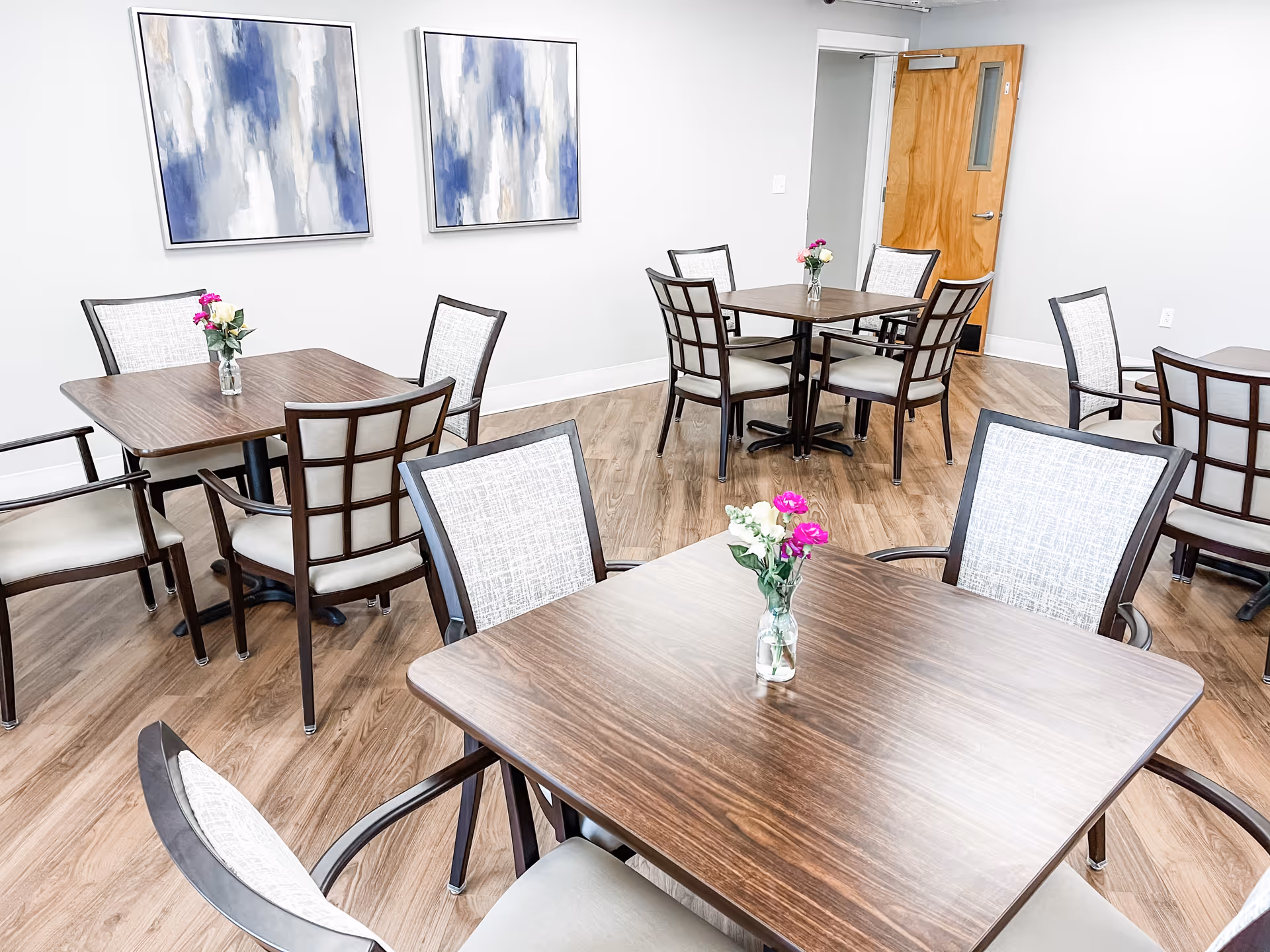 Bright communal dining room with wood tables and chairs, small vases of flowers on each table, and abstract artwork on the wall.