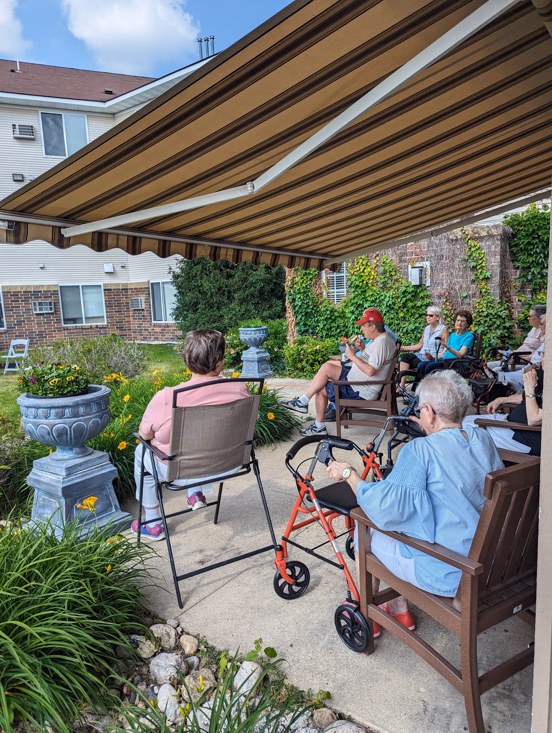 A group of elderly people sitting outdoors under a large striped awning at a senior living facility. They are seated on chairs arranged in a semi-circle on a concrete patio surrounded by greenery, flowers, and potted plants. Some individuals have walkers nearby. The building exterior with windows and brick accents is visible in the background.