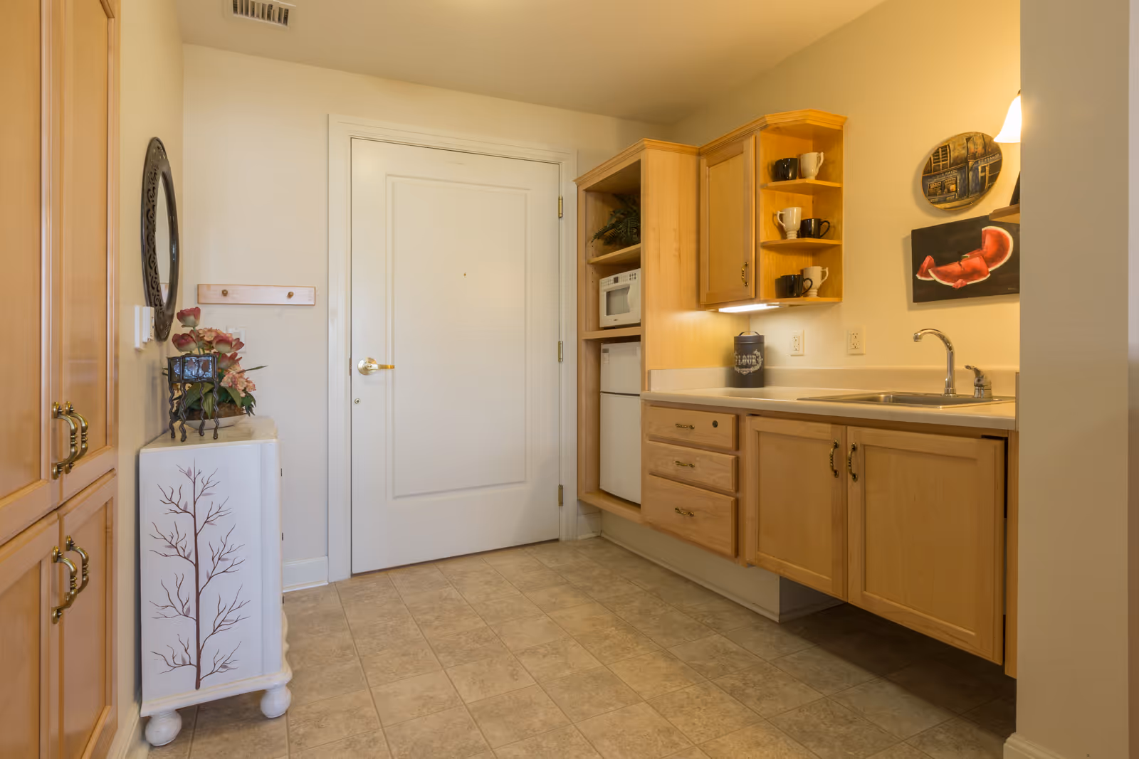 A small kitchen area with light wood cabinets, a countertop with a sink and faucet, a mini refrigerator, and a microwave. There is a white door in the background, a small decorative cabinet with a floral arrangement on top, and wall art including a round plate and a painting of a red lobster claw. The floor is tiled in a neutral color.