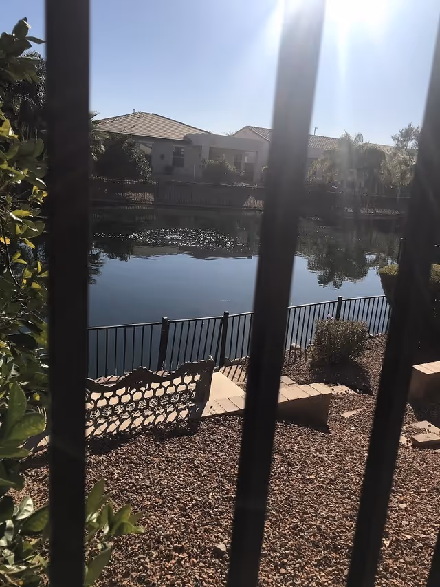 View through vertical bars of a fenced lakeside yard with a decorative bench, water, and houses across the pond under bright sun.