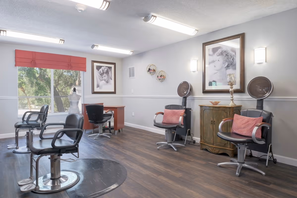 Interior view of a salon area with four black salon chairs, two hair drying stations, a wooden cabinet, framed black and white portraits on the walls, a window with a red valance, and a wooden floor.