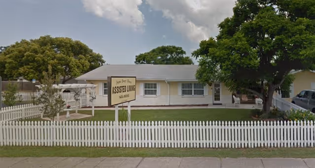 Single-story yellow assisted living home behind a white picket fence with a lawn and a sign reading 'Assisted Living'.